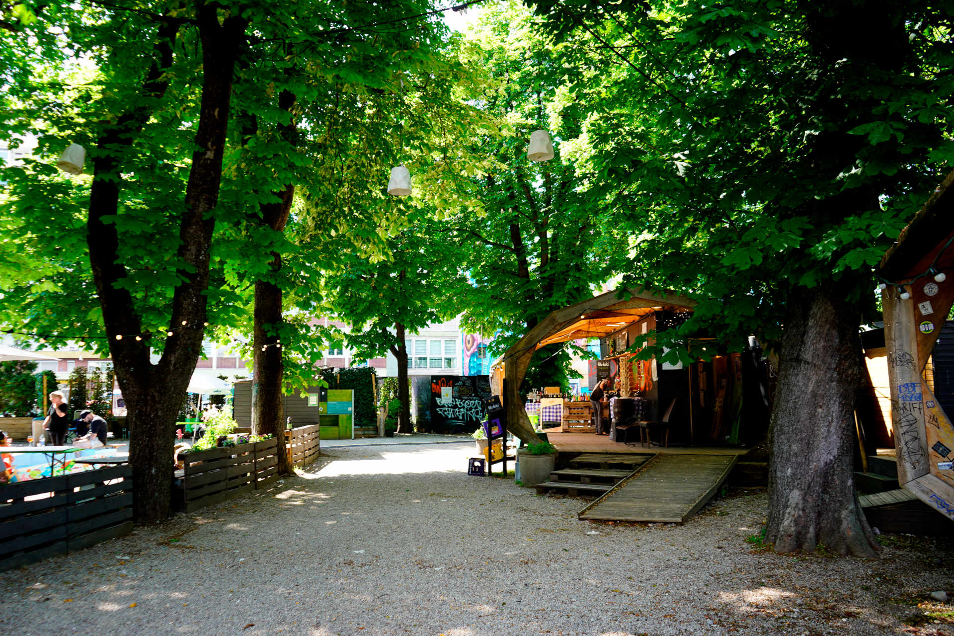 Une place avec de grands marronniers, du gravier en dessous, au fond à droite de l'image un pavillon en bois avec un kiosque.