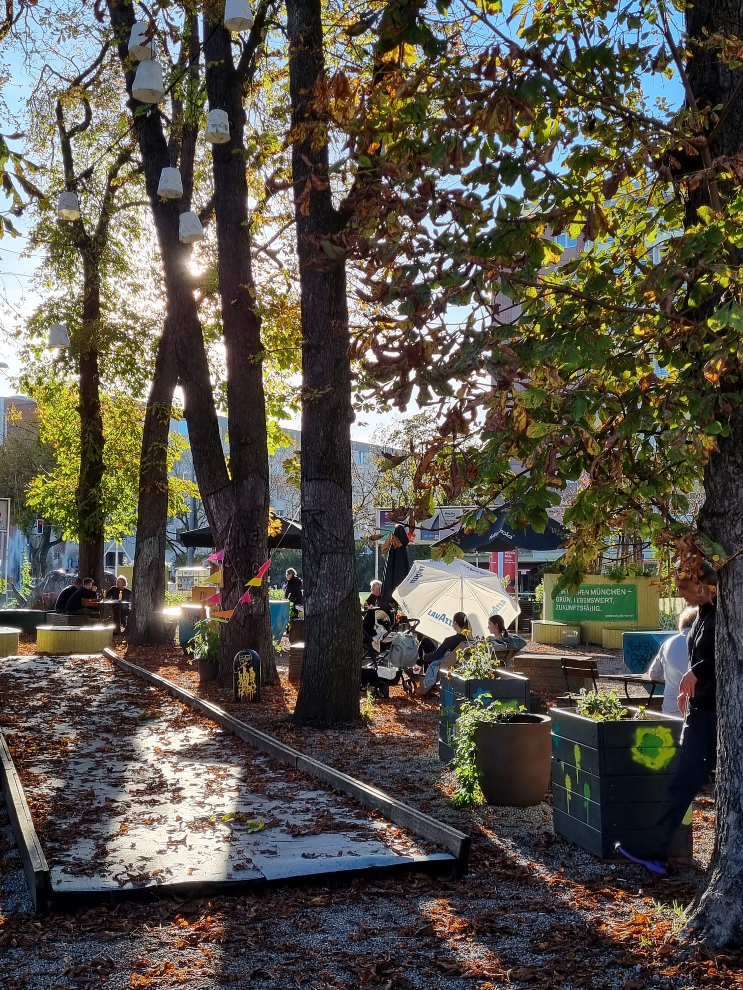Une place avec de grands marronniers, en dessous à gauche un terrain de pétanque recouvert de feuilles, à droite des parterres surélevés et des personnes assises à des tables de jardin.
