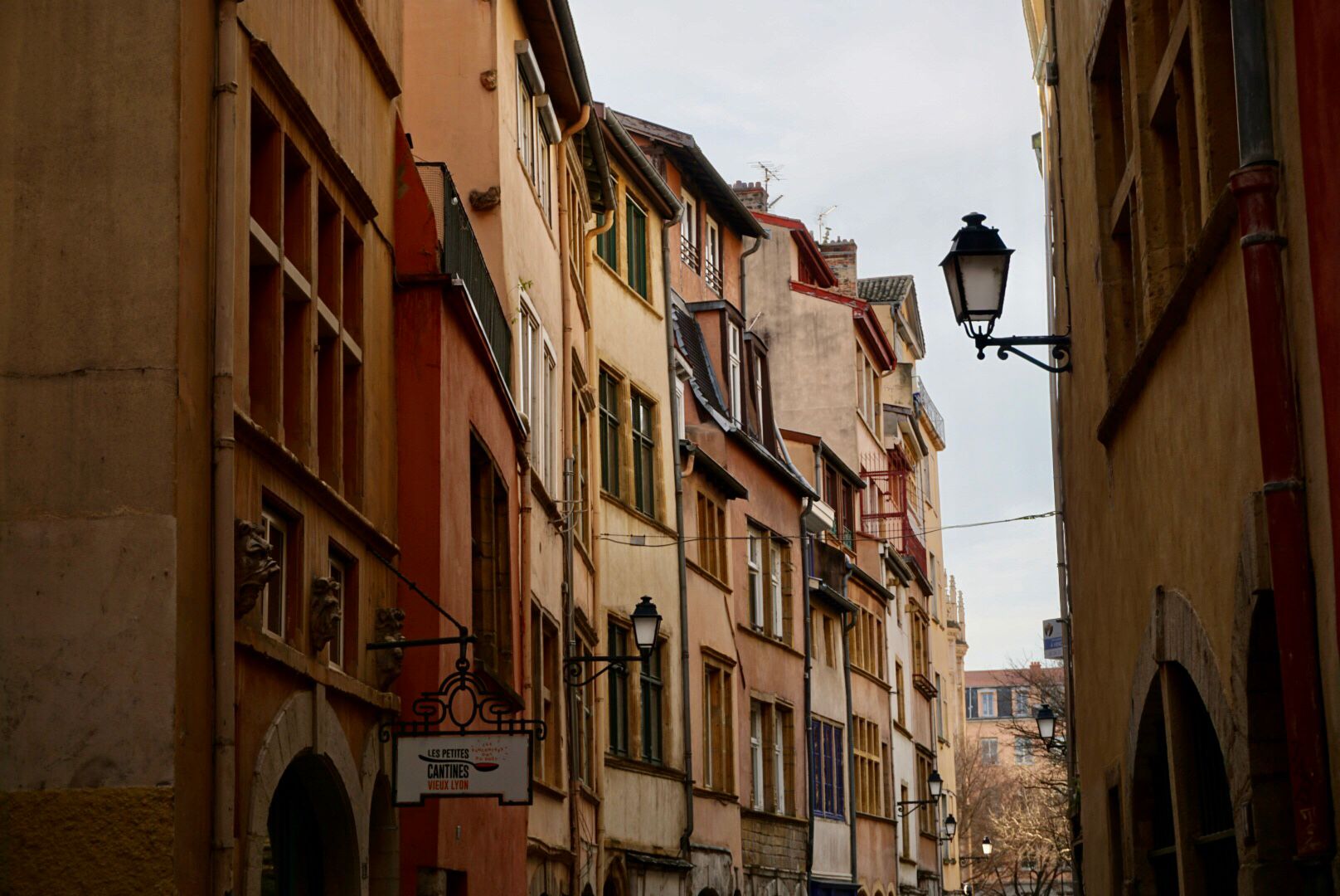 Rue étroite, bordée d'immeubles anciens à plusieurs étages et aux couleurs vives, avec des pavés et un lampadaire classique.