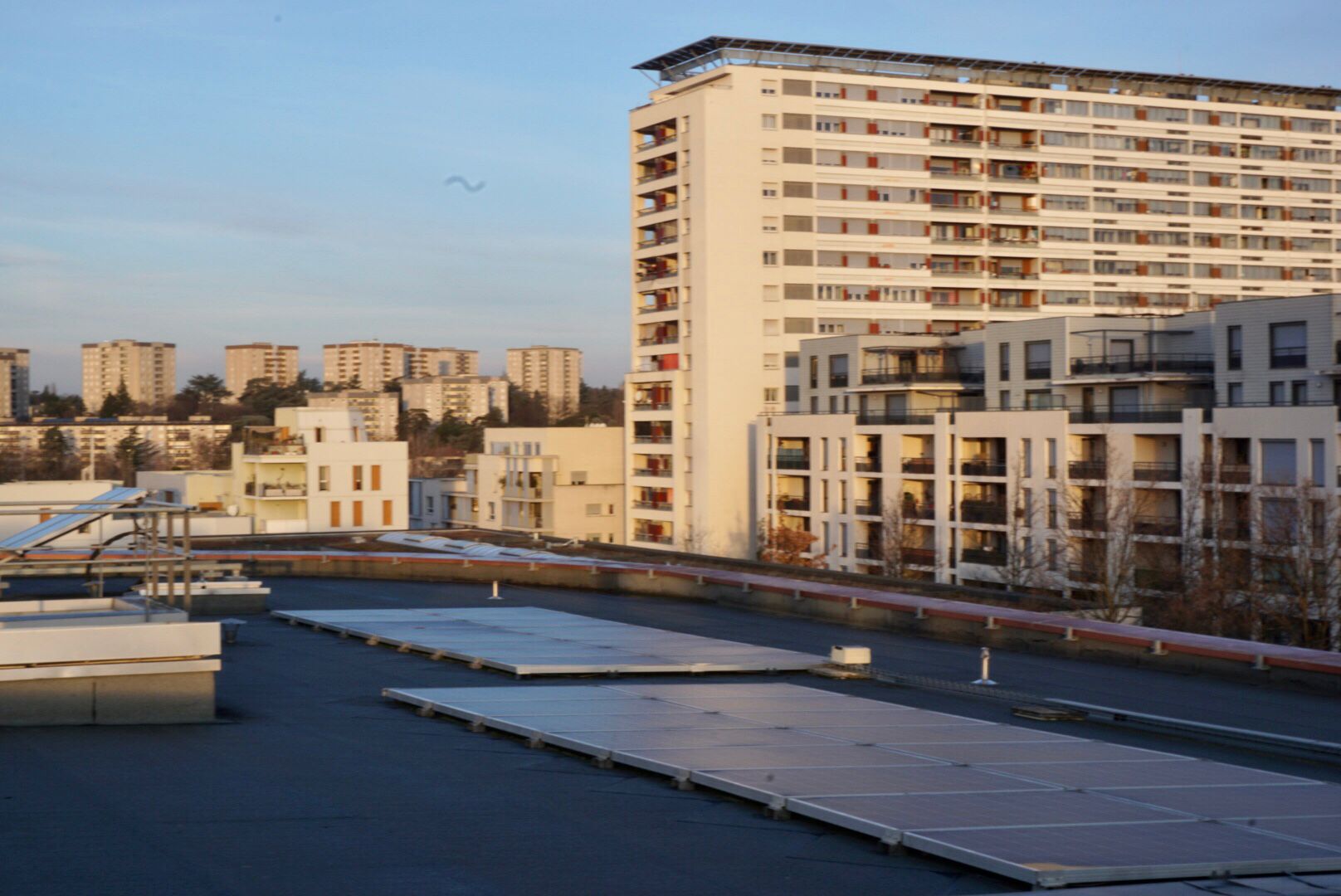 Vue d'un toit plat recouvert de panneaux solaires et, derrière, d'un quartier résidentiel dans la lumière du soir, avec des immeubles de grande hauteur.