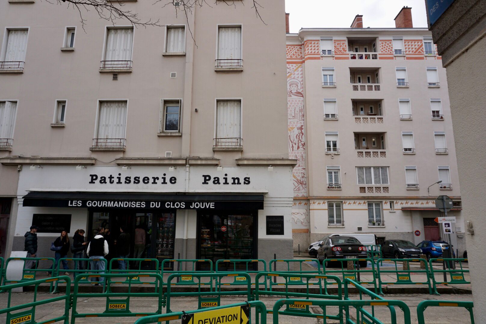 Des immeubles résidentiels de plusieurs étages avec des commerces au rez-de-chaussée, dont une boulangerie avec l'inscription "Pâtisserie Pains", derrière une clôture verte.