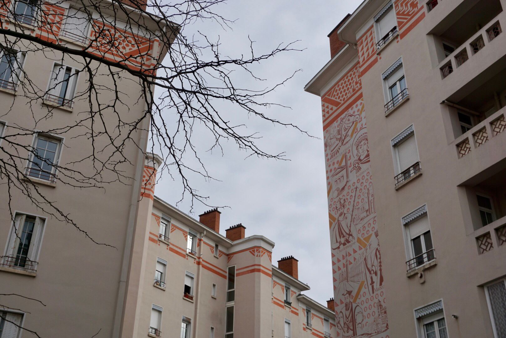 Des immeubles résidentiels de plusieurs étages avec des ornements rouge-orange et une grande fresque murale détaillée sur la façade, sont encadrée par un arbre nu.