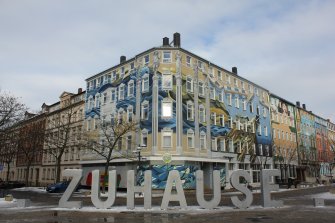 Une rue avec des façades de bâtiments artistiquement peintes en arrière-plan. Dans la rue, l'inscription « Home » est écrite en lettres géantes dans une tôle d'acier laquée.