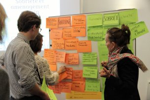 Trois personnes se tiennent devant un tableau d'affichage sur lequel sont collés de nombreux post-it. Sur les post-its, il y a des phrases sur la suffisance.