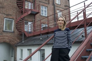 Une personne de genre féminin, avec des cheveux blonds mi-longs et un pull à rayures noires et blanches, se tient debout sur l’escalier devant un immeuble.