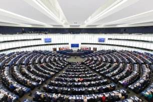 L'image montre l'hémicycle du Parlement européen à Strasbourg. La salle en demi-cercle est remplie de députés assistant à une séance. Au centre, une tribune avec le symbole de l'UE et des drapeaux des États membres. Au-dessus, des tribunes pour les spectateurs. L'architecture est moderne et lumineuse.