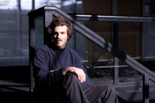 Une personne vêtue de noir, aux cheveux courts et foncés, avec une moustache, est assise sur un escalier.